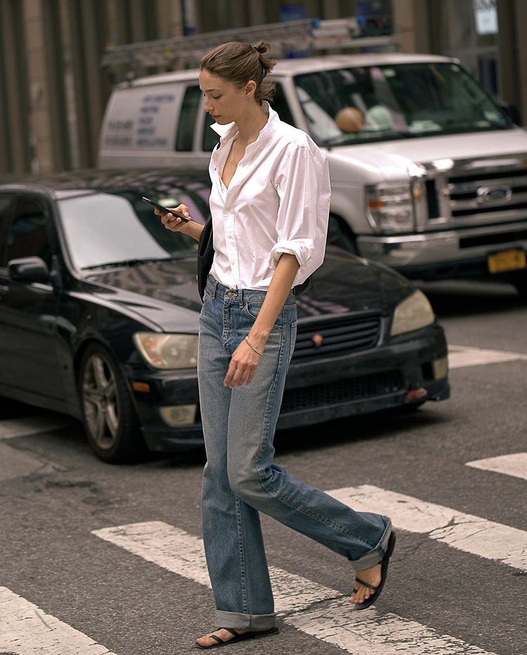 Woman in a white button-down shirt, straight jeans, and black sandals walking across a city street