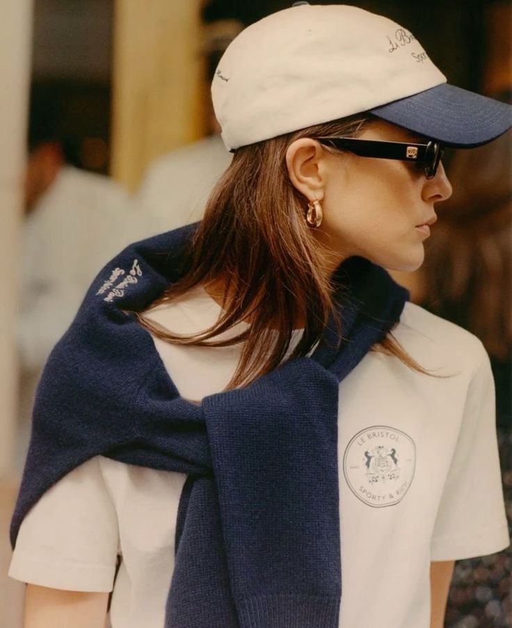 Woman in a branded cap, sunglasses, and sweater draped over a tee showing how quiet luxury can become a recognizable code instead of a resolved look