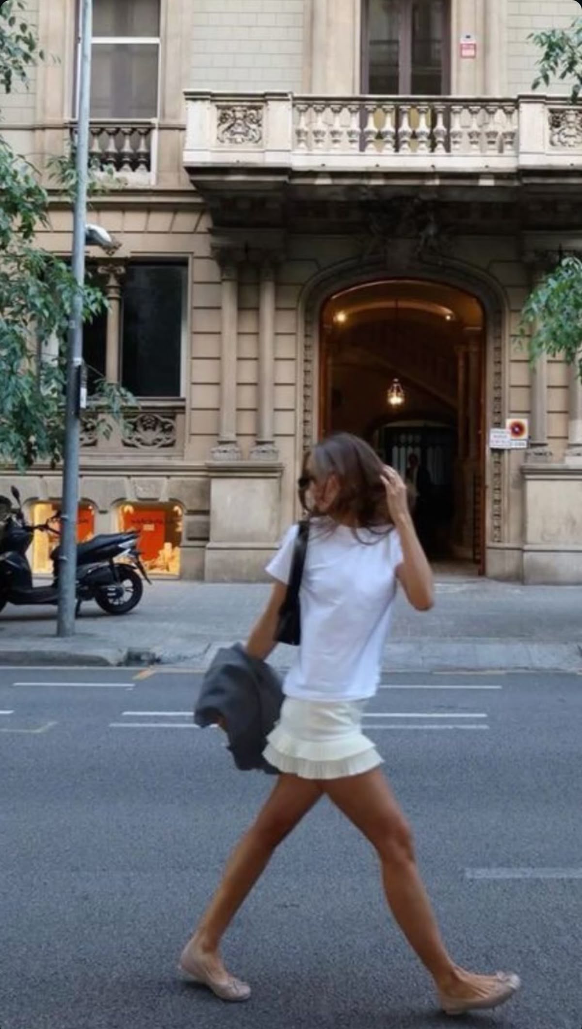 Woman in a white tee and short white skirt walking past a stone building with a soft, fleeting off-duty ease