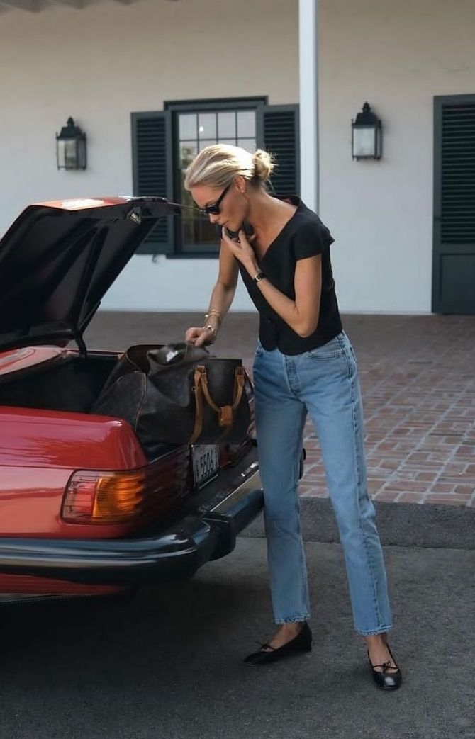 Woman in a black top, straight jeans, and flats beside a red car showing denim as connective tissue in the outfit