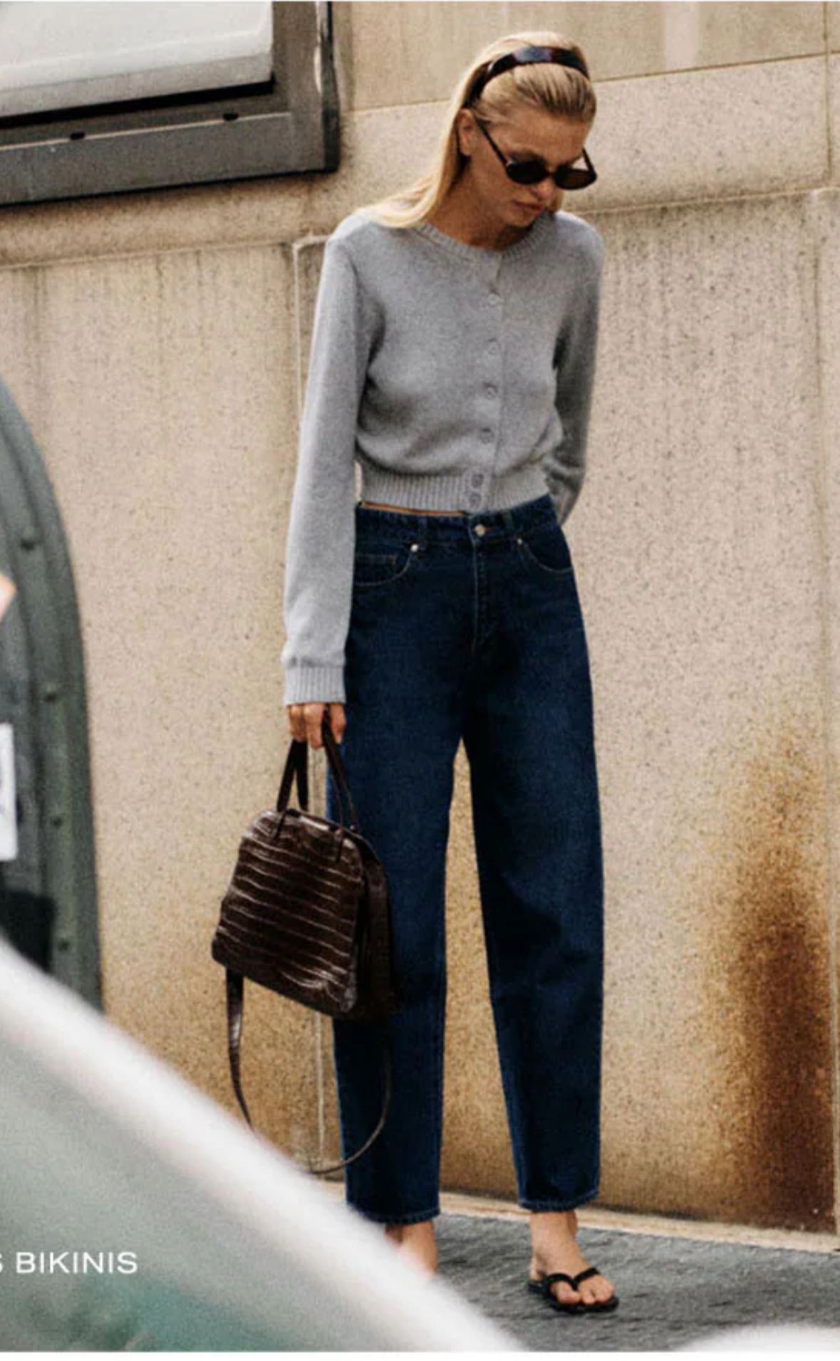 Woman in a grey cardigan, dark jeans, sunglasses, and a structured handbag
