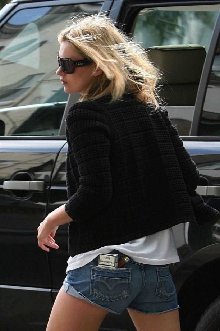 Woman in sunglasses, a dark cardigan, and denim shorts turning away beside a black car